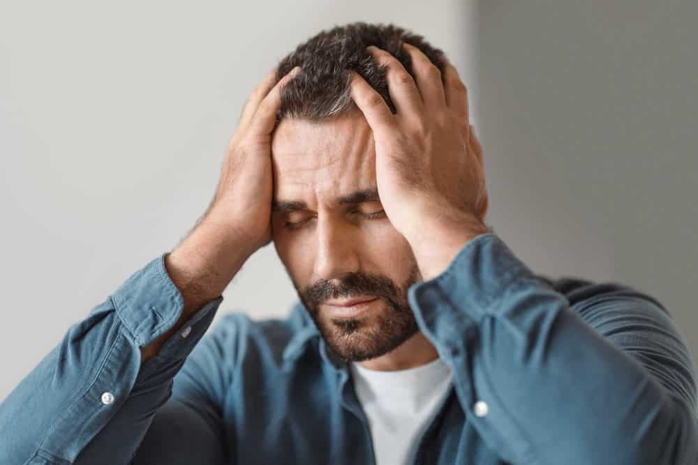 Man in a blue shirt holding his head with both hands, eyes closed, appearing stressed or frustrated.