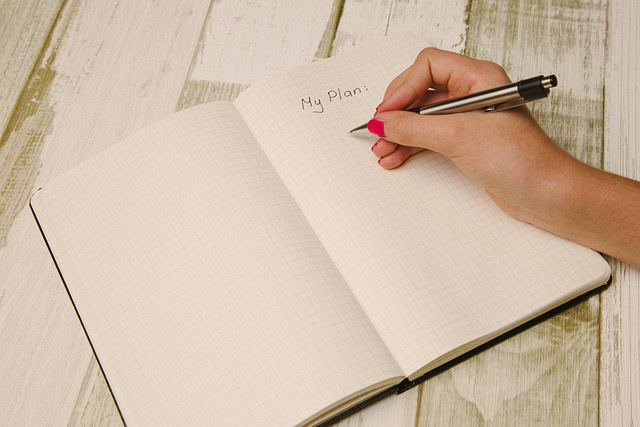 Hand writing My Plan at the top of a graph paper notebook with a pen, on a wooden surface.