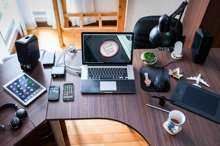 A workspace with a laptop displaying Start Engine Stop, surrounded by various devices such as headphones, a tablet, phones, and a graphics tablet; all enveloped in the subtle hum of wifi radiation. Nearby sit a cup of tea, plant, model airplane, and lamp.