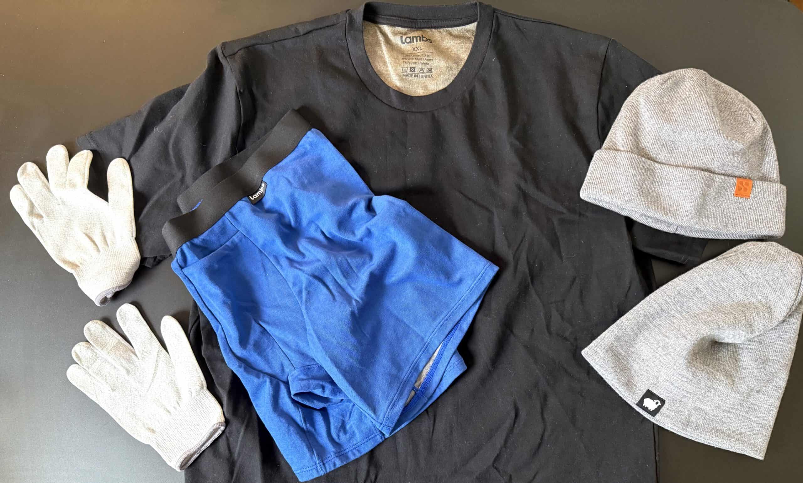 Flat lay of clothing items on a black surface: a black T-shirt, blue underwear, white gloves, and two gray beanies.