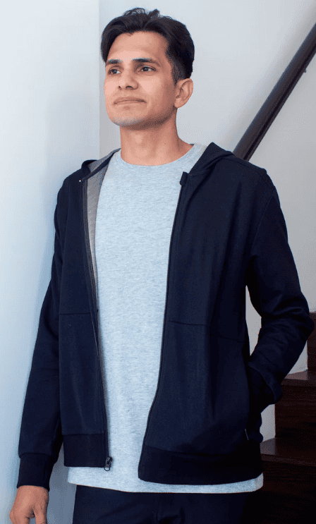 Person wearing a gray shirt and black hoodie, standing near a staircase, with one hand in a pocket, looking forward.