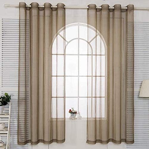 Light brown sheer curtains hang over a large arched window with a white frame. A small plant sits on the windowsill, and a lamp is partially visible on the right.