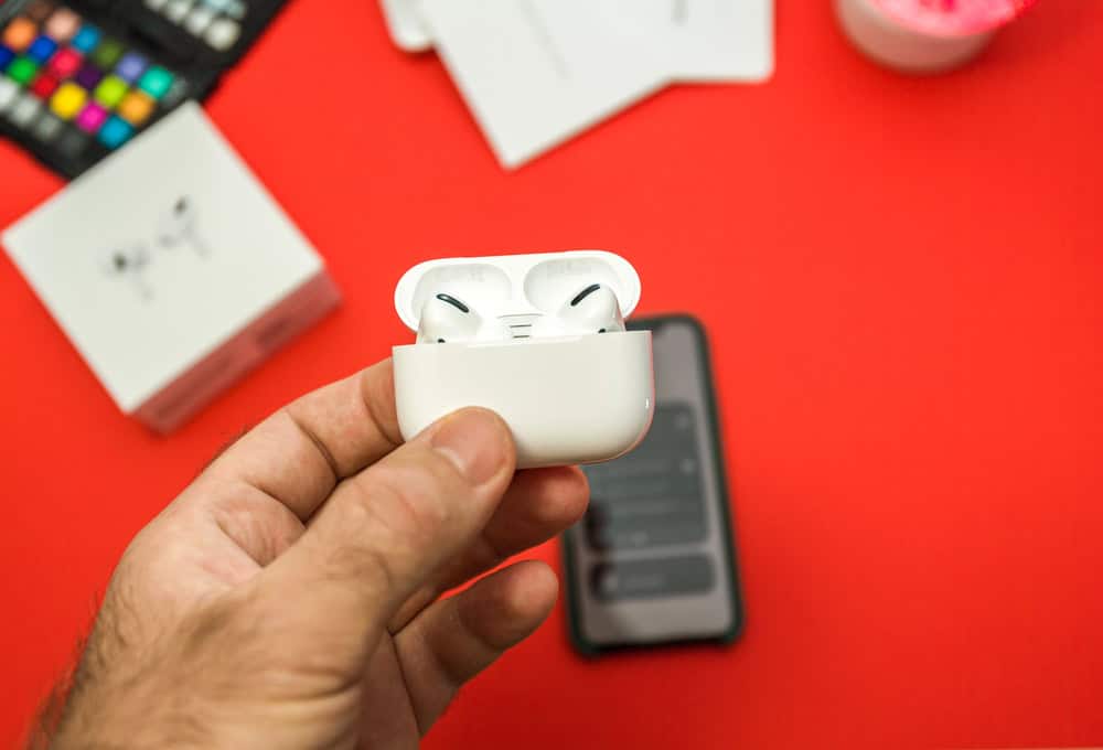 A hand holds an open AirPods Pro case with earbuds, with an iPhone, AirPods box, and other items blurred in the background on a red surface.