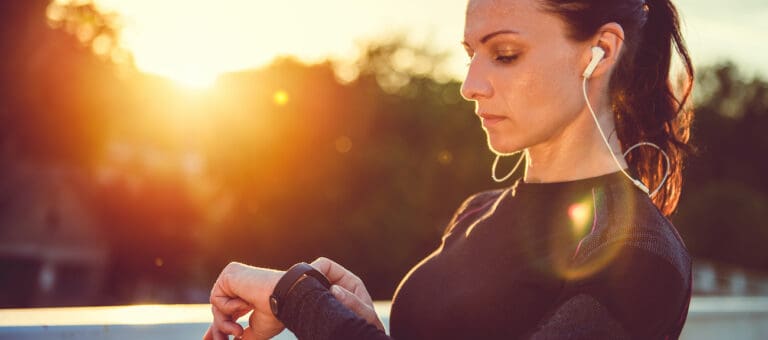 A woman in athletic wear checks her smartwatch while wearing earphones during a sunset.