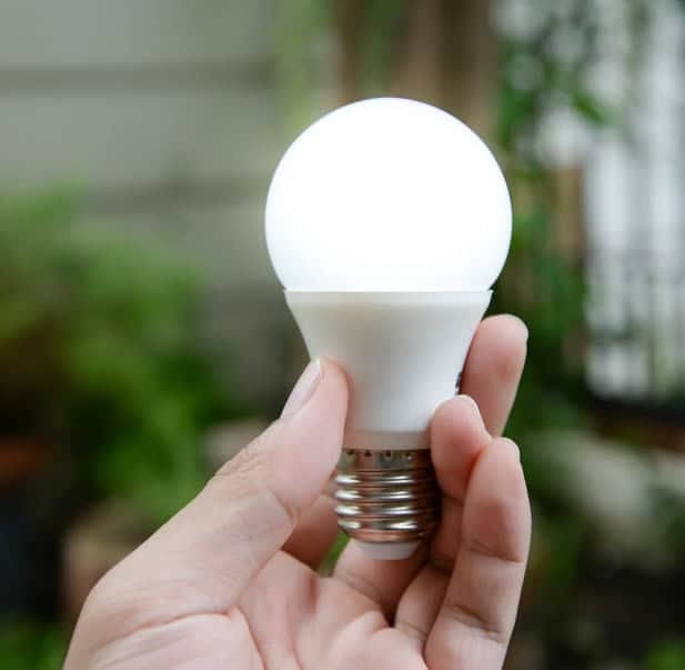 A hand holding a LED light bulb outdoors, with a blurred green background.