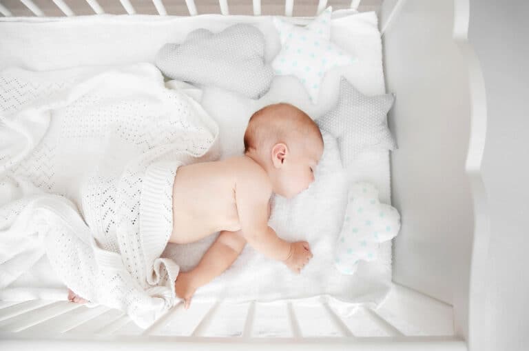 A baby sleeps on their stomach in a crib, covered with a white blanket. The crib contains star and cloud-shaped pillows.