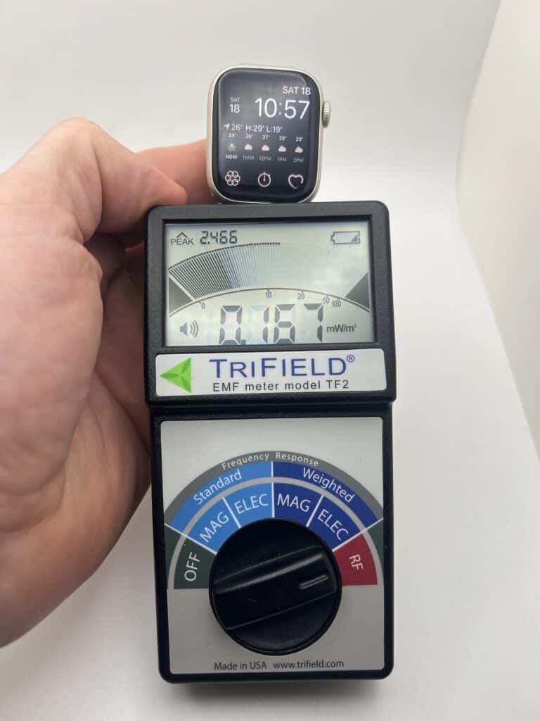 A person holds an Apple Watch above a TriField EMF meter displaying a reading of 0.876 mW/m². The watch shows date and time as Saturday 18th, 10:57.