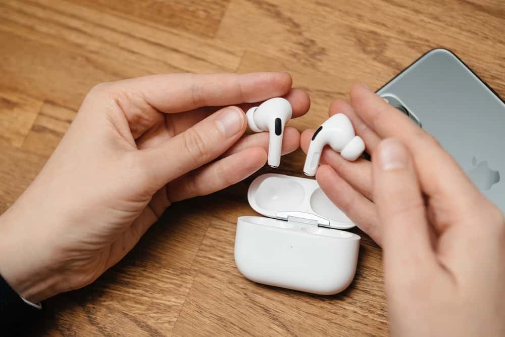 Hands holding wireless earbuds above an open charging case on a wooden surface, with a smartphone nearby.