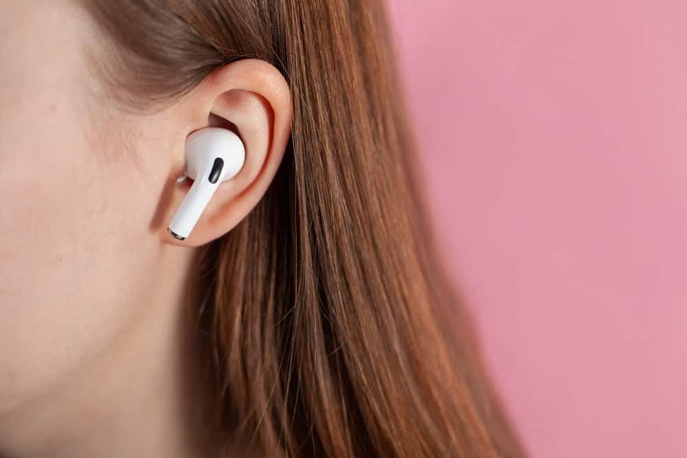 Person with long brown hair wearing a white wireless earbud in their right ear against a pink background.