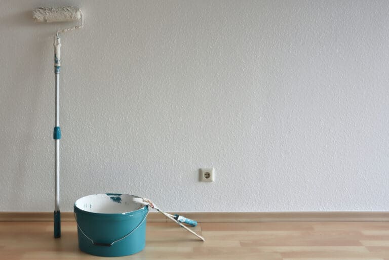A partially painted wall with a paint roller, a blue bucket of white paint, and a brush on a wooden floor.
