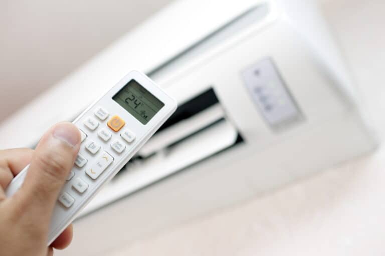 A hand holds an air conditioner remote set to 24°C, pointing towards a wall-mounted air conditioning unit.