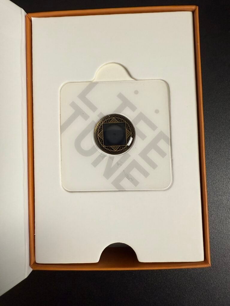 A geometric black ring with a hexagonal design is displayed in an open box with white interior and text partially visible underneath.