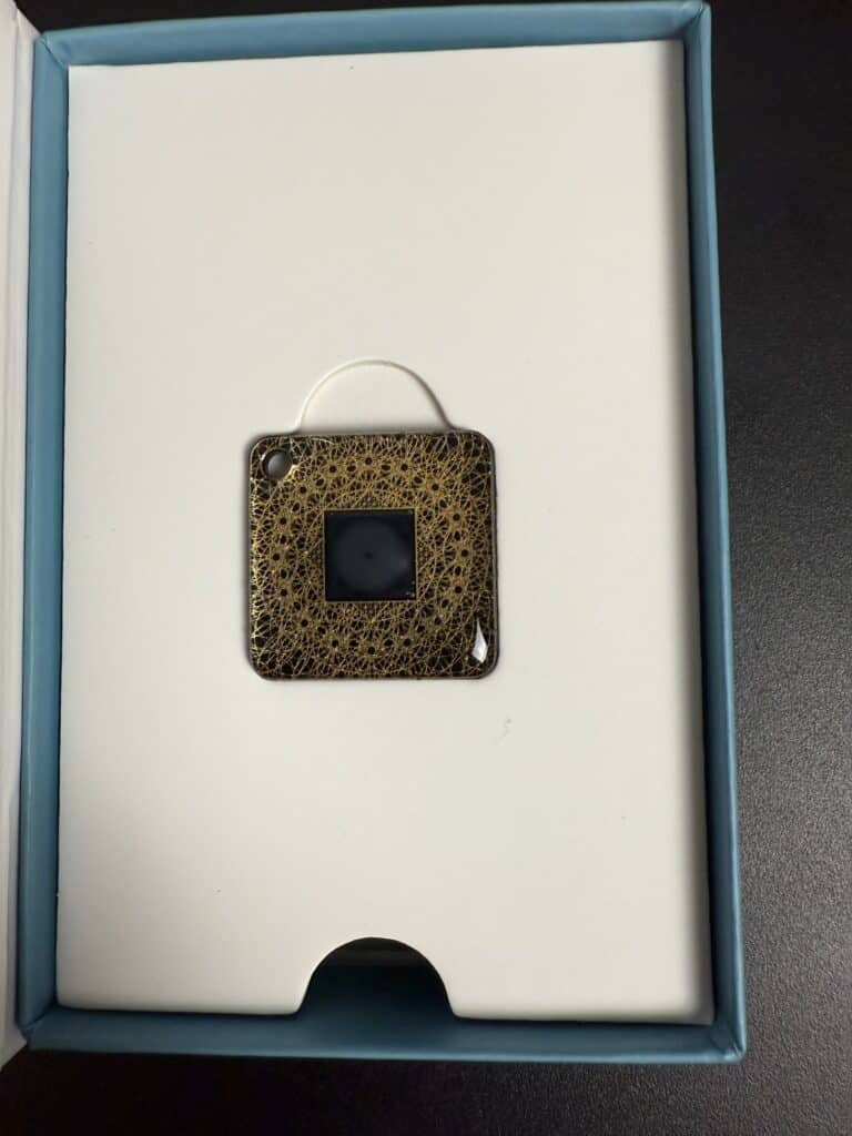 A square, decorative electronic device with a small circular hole at the corner, set in a white box on a black surface.
