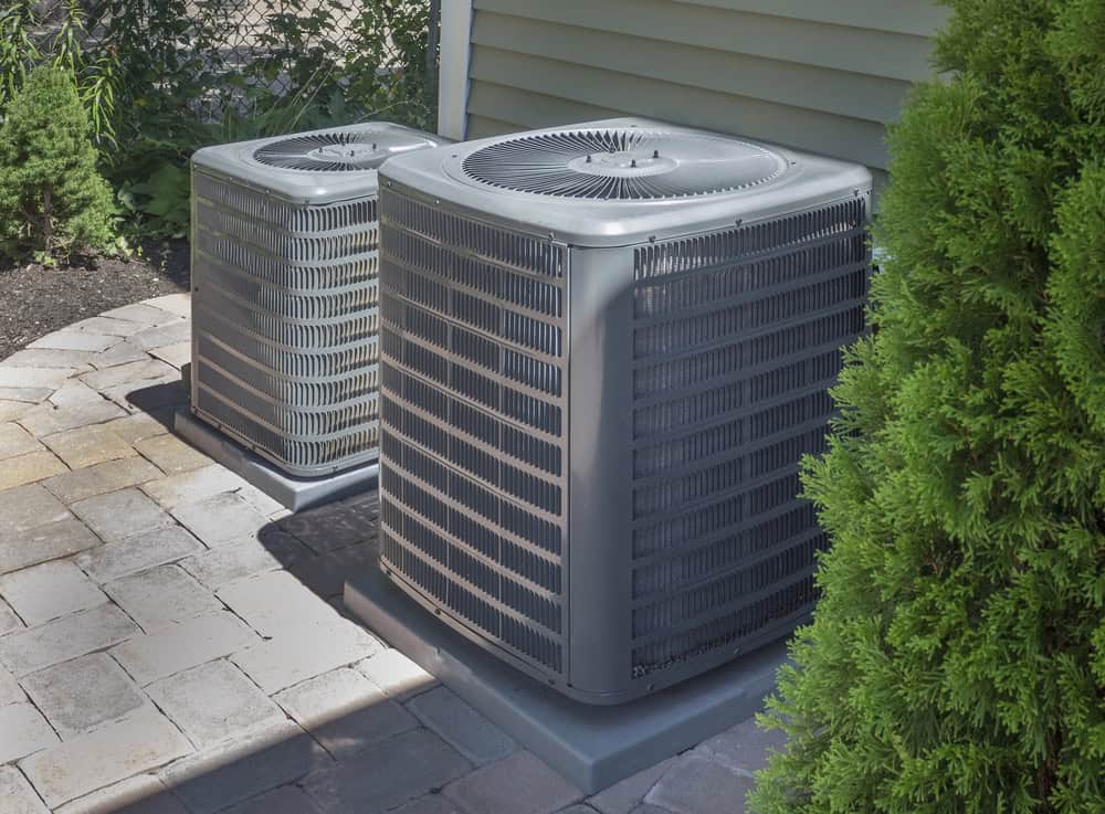 Two outdoor air conditioning units on a paved patio beside a house, surrounded by greenery.