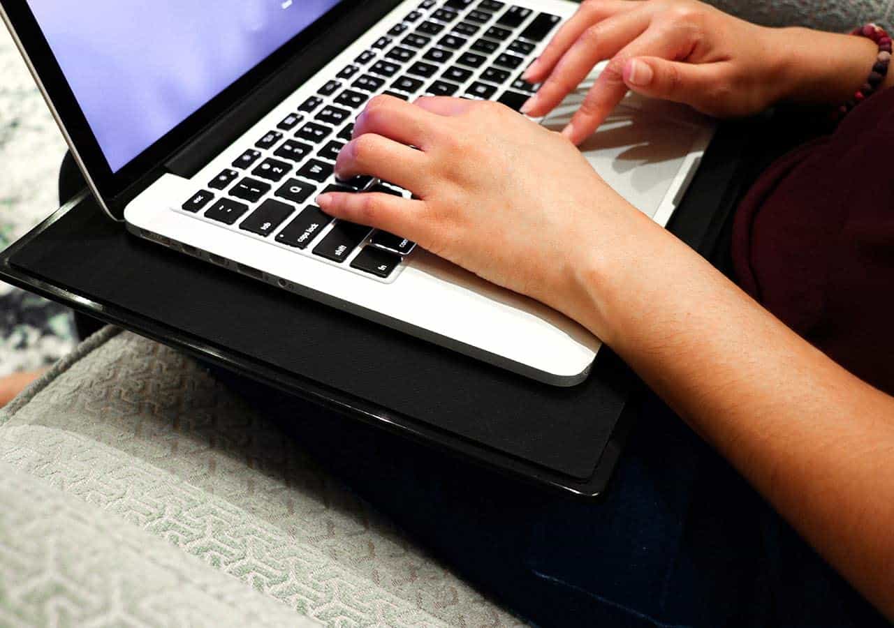 A person typing on a laptop placed on their lap, sitting on a cushioned chair.