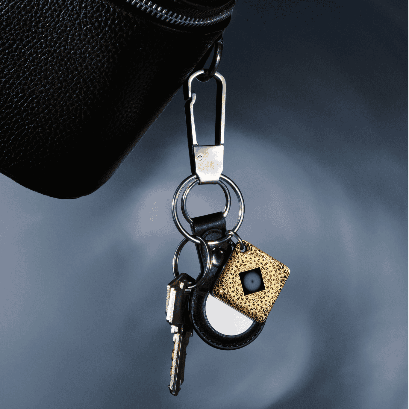 Screenshot 2026-01-06 at 93549 PM - EMF Academy A set of keys, a round black-and-white fob, and a gold square tracker are attached to a black bag with a metal carabiner clip against a blurred background.