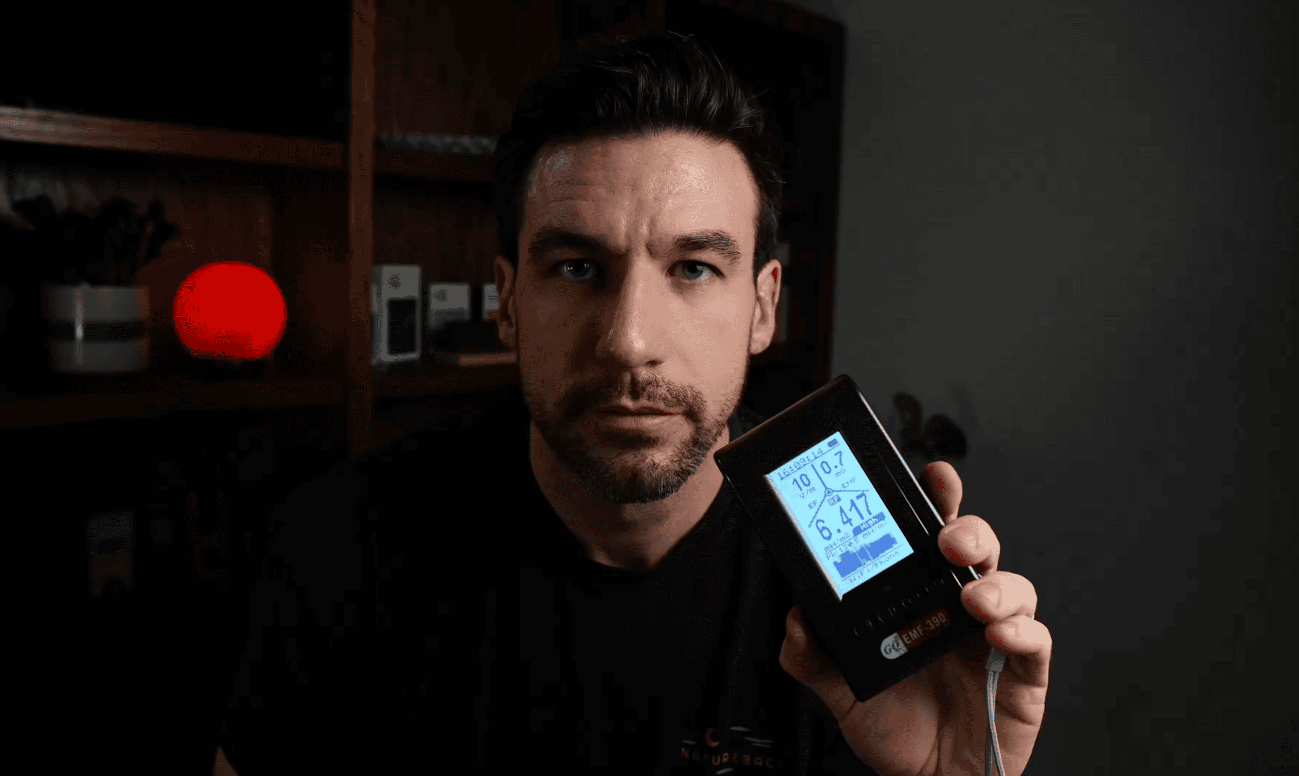 A man sits indoors holding up a digital device with a screen displaying various readings, including numbers and icons. Shelves with objects are visible in the background.