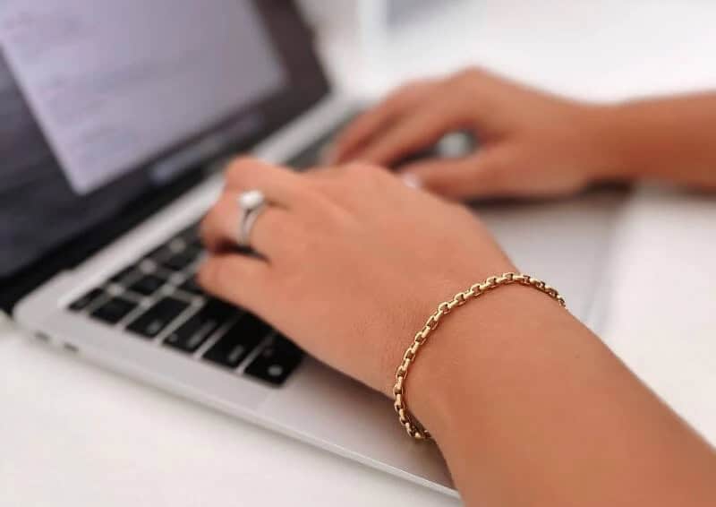 Hands typing on a laptop keyboard, wearing a silver ring and a gold chain bracelet.