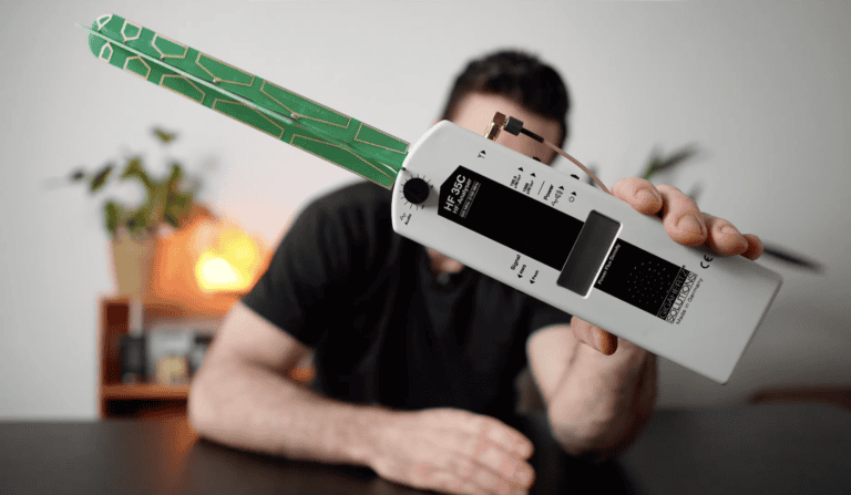 A person holds a white electromagnetic field (EMF) meter with a green circuit board antenna in front of their face, seated at a black table.