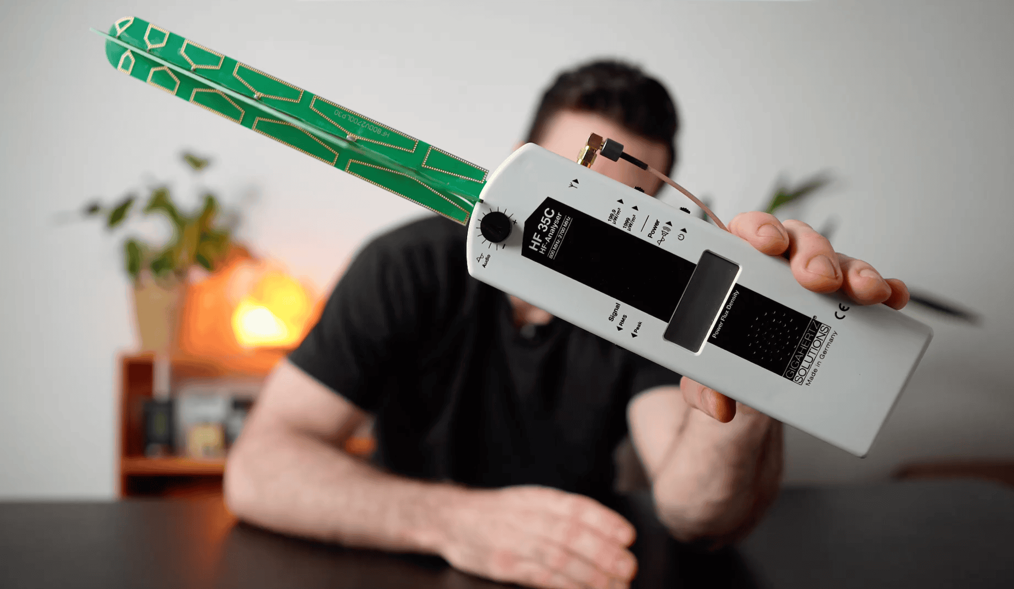 A person holds a white electromagnetic field (EMF) meter with a green circuit board antenna in front of their face, seated at a black table.