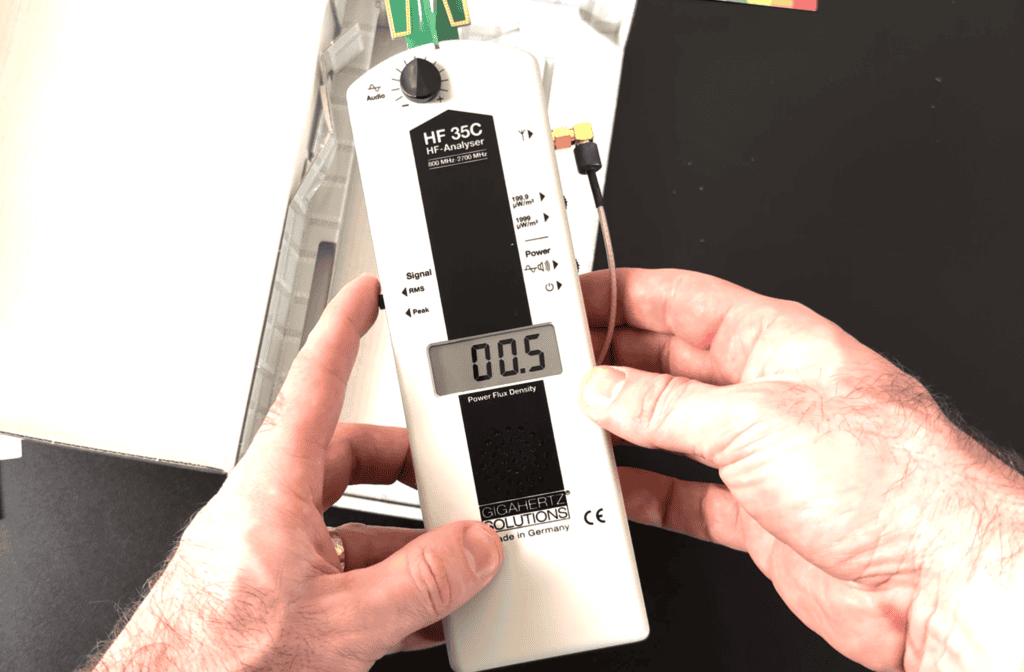 Screenshot 2026-02-11 at 112204 AM - EMF Academy A person holds an HF-35C RF analyzer showing a power flux density reading of 0.5 on its digital display.