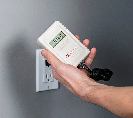 A person holds an EMF meter displaying a reading of 0.43 near an electrical outlet.