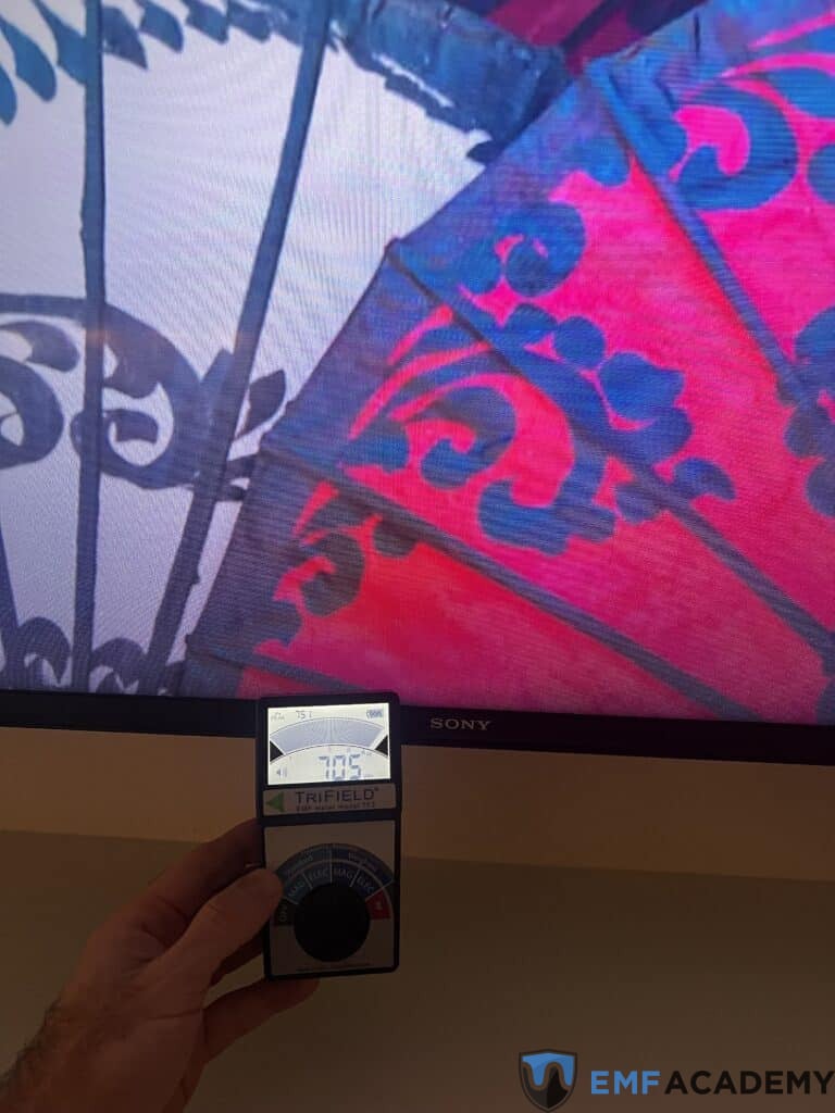 A hand holds a Trifield meter showing a reading of 3.75 in front of a tv screen.