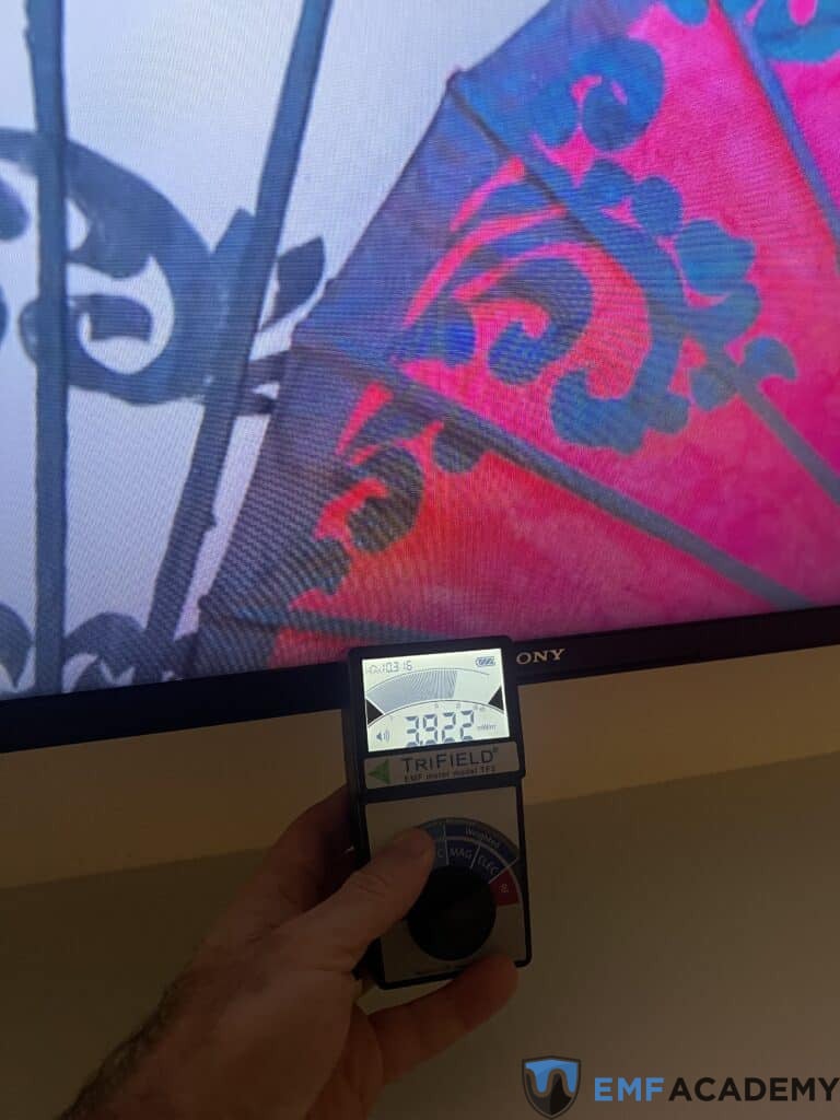 A hand holds a Trifield meter displaying a reading of 383.5 in front of a TV screen.