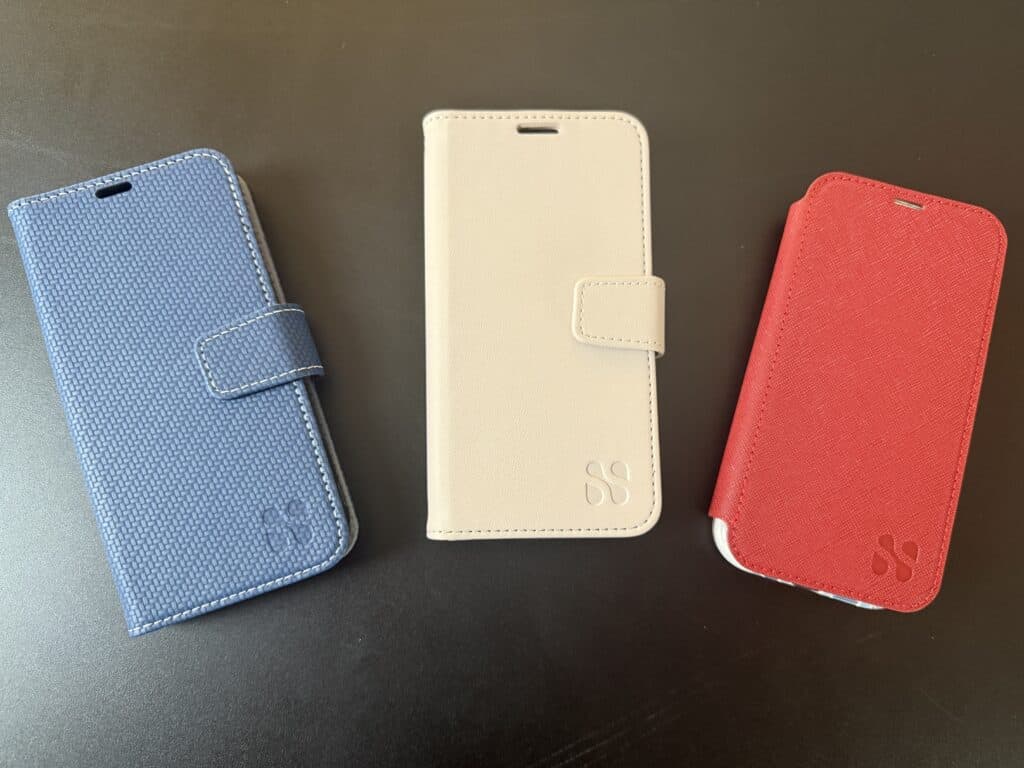 Three smartphone wallet cases in blue, white, and red are displayed on a black surface, each with a clasp and a subtle logo imprint.
