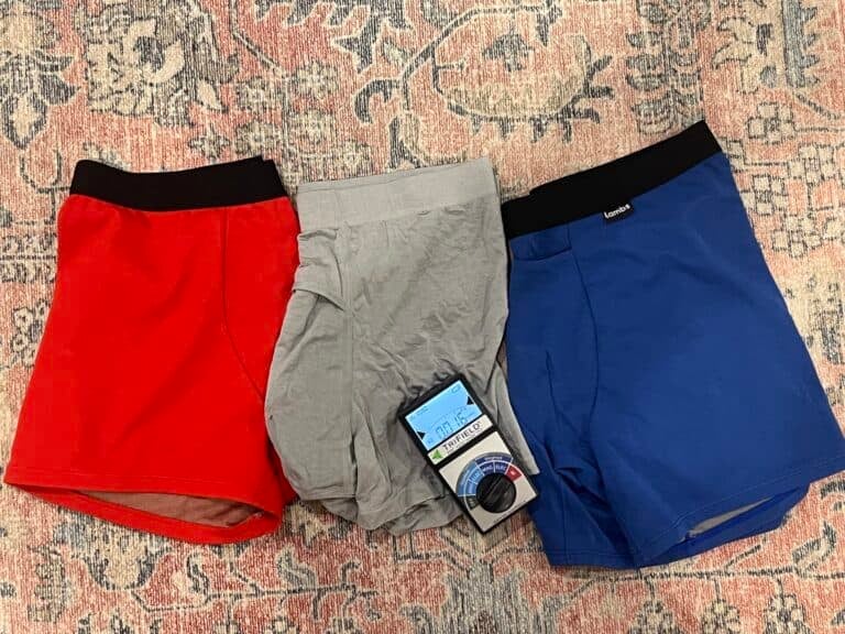 Three pairs of mens boxer briefs in red, gray, and blue are laid out on a patterned fabric background. The gray pair has a label attached.