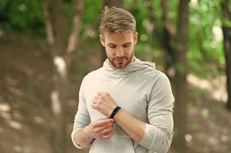 A man in a light gray hoodie looks at a fitness tracker on his wrist while standing in a wooded area.