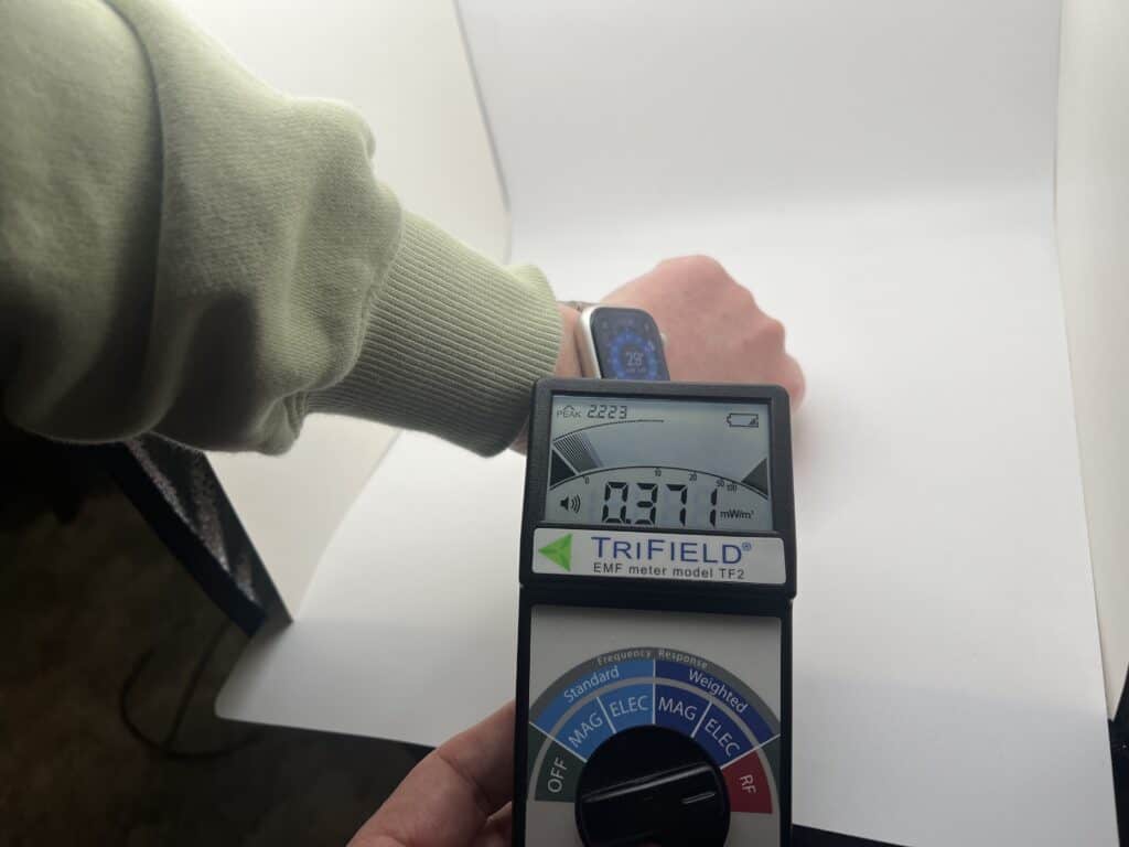 A person holds a Trifield EMF meter near a smartwatch on a wrist, displaying a reading of 0.37.