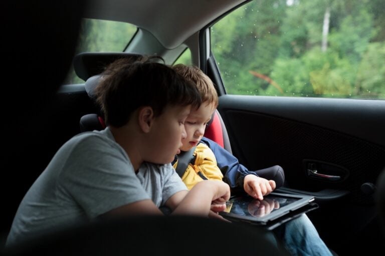 Two young boys sit in the backseat of a car, closely looking at and using a tablet together, with trees visible outside the window.