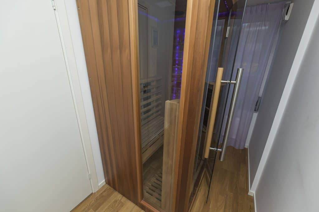 A modern wooden sauna with glass door and metal handle, located indoors with wooden flooring and white walls.