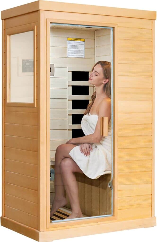 A woman wearing a towel sits inside a wooden infrared sauna with a glass door and visible control panel.