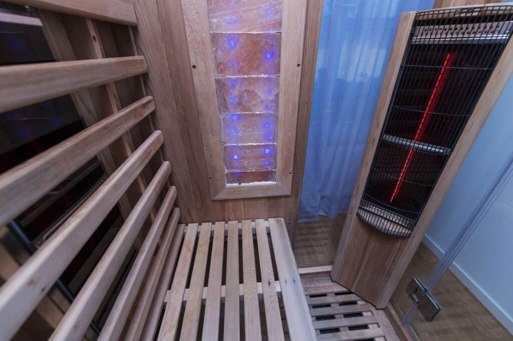 Wooden infrared sauna interior with a slatted bench, Himalayan salt panel with blue lights, and an electric heater, next to a glass door and sheer curtain.