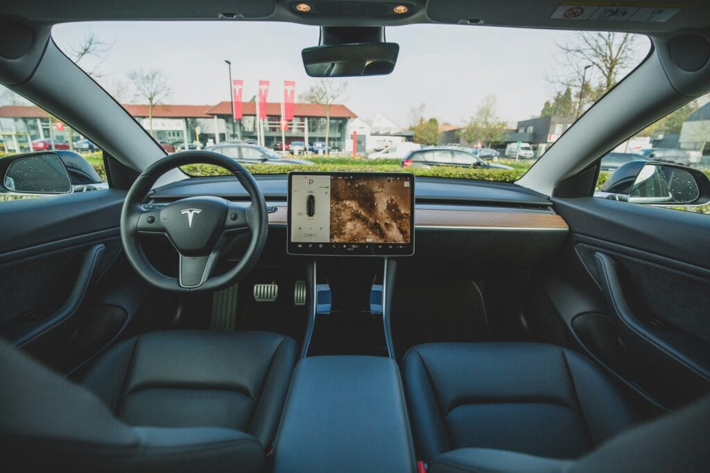 EMF EV Radiatoin image - EMF Academy Interior view of a Tesla car showing the steering wheel, touchscreen display with maps, and black seats, taken from the back seat during daytime.