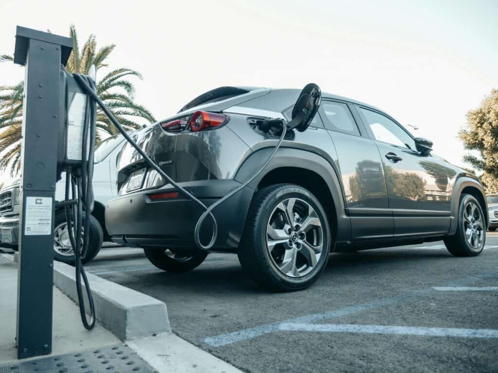 A gray electric car is parked and plugged into a charging station in an outdoor lot on a sunny day.