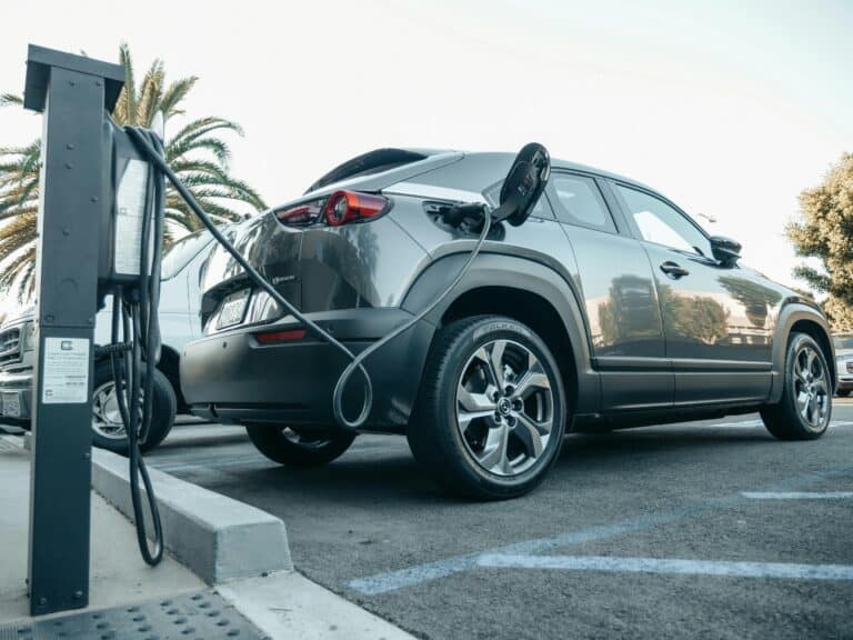 A gray electric car is parked and plugged into a charging station in an outdoor lot on a sunny day.