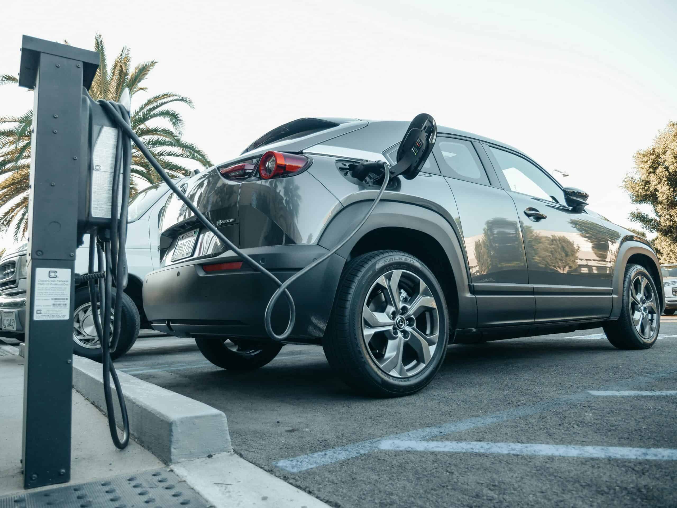 A gray electric car is parked and plugged into a charging station in an outdoor lot on a sunny day.