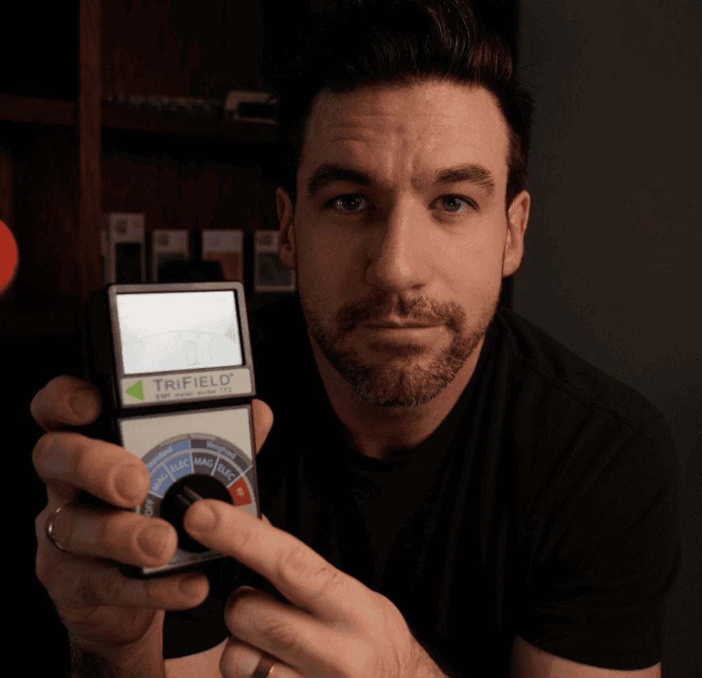 A man holds up a TriField meter, pointing at its dial, with shelves and a glowing orange object in the background.