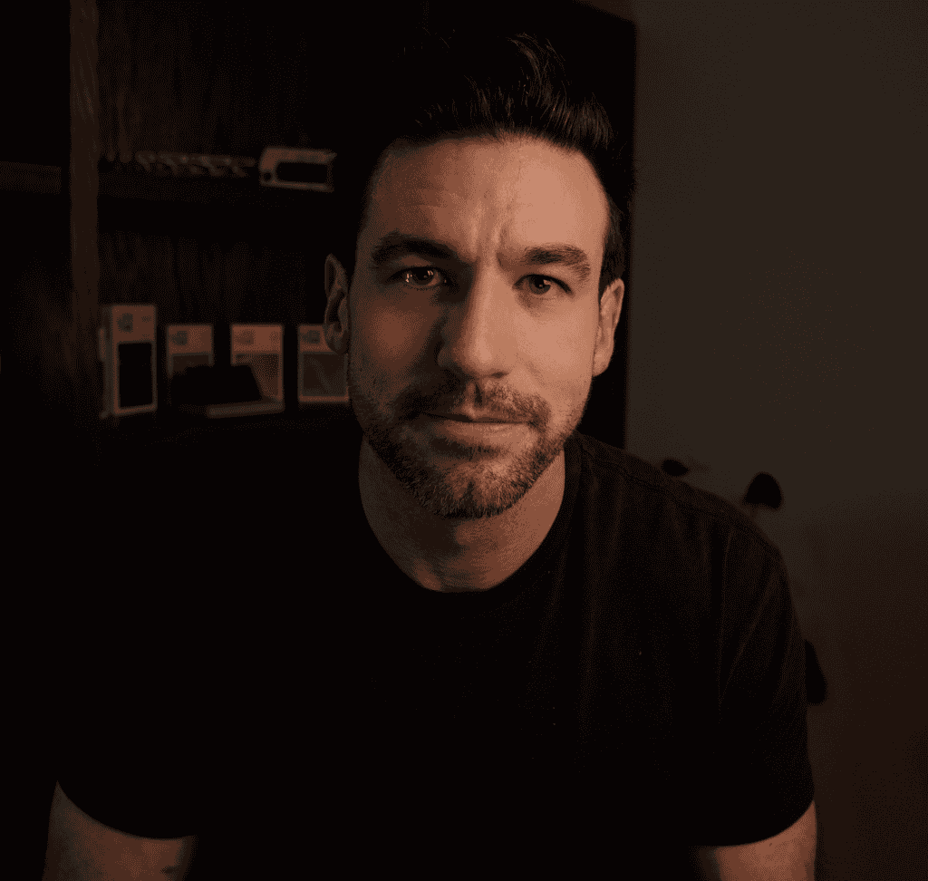 Screenshot 2026-02-16 at 50654 PM - EMF Academy A man with short dark hair and a trimmed beard, wearing a black t-shirt, sits indoors in front of a wooden shelf with small boxes on it.